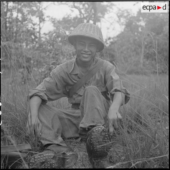 22 Militaire cambodgien assis par terre, chaussé de brodequins cloutés et coiffé d'un casque viêt minh abandonné sur le [description provisoire]