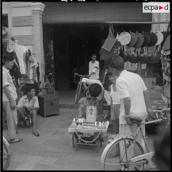 17 Dentiste ambulant effectuant des soins sur un enfant, dans la rue près d'un présentoir de bérets et de chapeaux. [description provisoire]