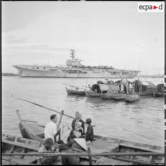 Arrivée du porte-avions Arromanches dans le port de Saigon où mouillent des jonques et des sampans.