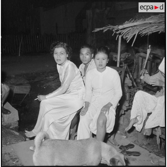La vie nocturne à Saigon : un homme assis dans la rue auprès de deux femmes en tenue traditionnelle (Ao dai).