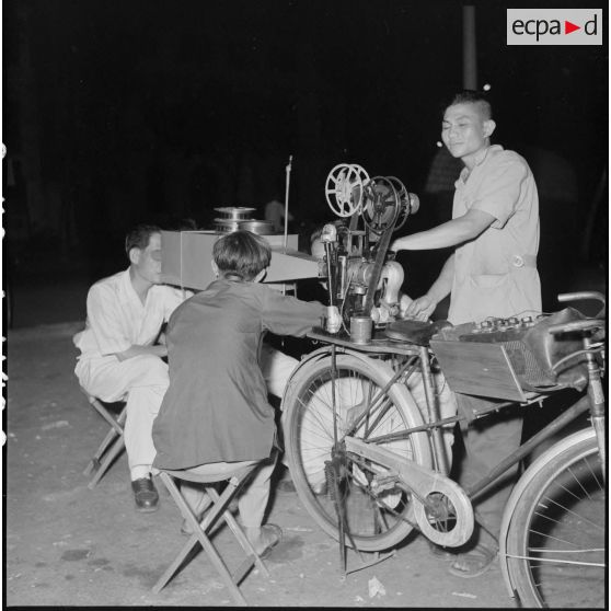 Dans une rue de Saigon, visionnage d'un film à partir d'un appareil de projection transformé en visionneuse et installé à l'arrière d'une bicyclette.