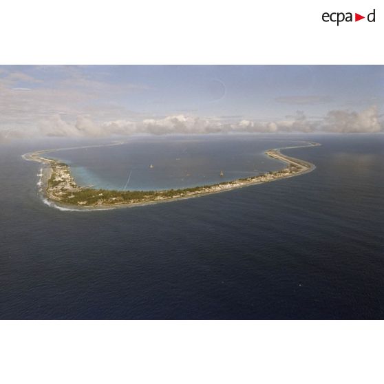 Vue aérienne de l'atoll de Moruroa côté océan (au point kilométrique PK 18) avec un fond de ciel nuageux. Au premier plan, la zone de vie du centre d'expérimentations du pacifique (CEP), et la piste de l'aéroport. Dans le lagon, la plateforme de forage Tila.