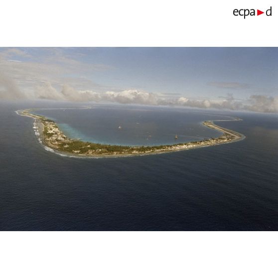 Vue aérienne de l'atoll de Moruroa côté océan (au point kilométrique PK 18) avec un fond de ciel nuageux. Au premier plan, la zone de vie du centre d'expérimentations du pacifique (CEP), et la piste de l'aéroport. Dans le lagon, la plateforme de forage Tila.