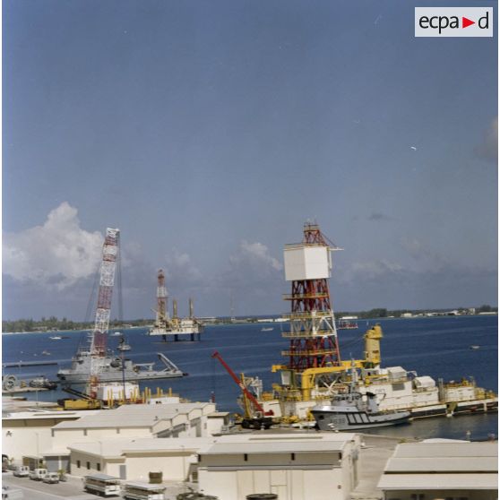 Vue aérienne des barges du Commissariat à l’énergie atomique (CEA) à Moruroa : le remorqueur portuaire Manini (A638) près de la barge de forage Manutéré et une barge de forage à quai au port , et la plateforme autoélévatrice de forage de puits de tir Tila dans le lagon de l'atoll.