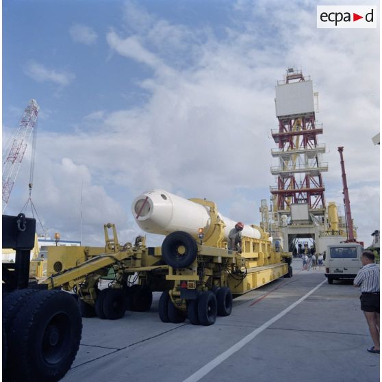 Le cylindre conteneur de l'engin destiné à l'essai du tir nucléaire est acheminé par un camion de transport lourd sur un quai du port de l'atoll de Morurua. La barge Manutéré de forage du Commissariat à l’énergie atomique (CEA) est en arrière-plan.