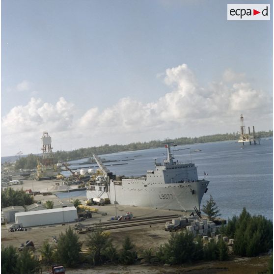 Vue aérienne du bâtiment de transport et de soutien (BTS) Bougainville (L9077) à quai dans le port de Moruroa devant la barge Manutéré de forage du Commissariat à l’énergie atomique (CEA). En arrière-plan, la plateforme autoélévatrice de forage de puits de tir Tila dans le lagon.
