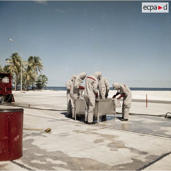 Sur une aire du service de décontamination de Hao, près d'une motopompe à eau à compresseur, des personnels en tenue de protection décontaminent du matériel dans un bac de lavage après un tir d'essai nucléaire.