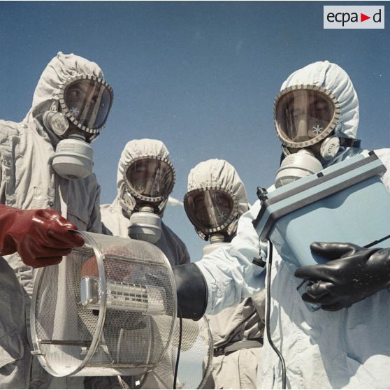 Sur une aire du service de décontamination de Hao, des personnels en tenue de protection testent la radioactivité de matériel avec un détecteur après un tir d'essai nucléaire.