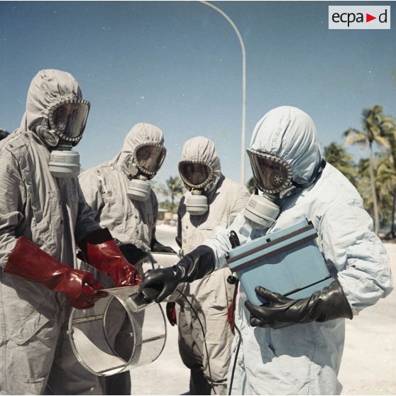 Sur une aire du service de décontamination de Hao, des personnels en tenue de protection testent la radioactivité de matériel avec un détecteur après un tir d'essai nucléaire.