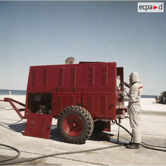 Sur une aire de lavage du service de décontamination de Hao, utilisation d'une motopompe à eau à compresseur par un personnel en tenue de protection pour décontaminer du matériel après un tir d'essai nucléaire.