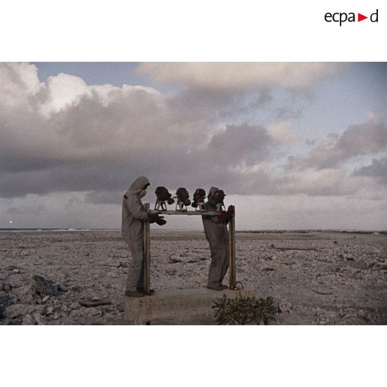 Près du poste d’enregistrement avancé (PEA) Denise, des personnels en tenue NBC relèvent des masques à gaz exposés pour étude des effets des tirs par la section technique de l’armée de terre (STAT).