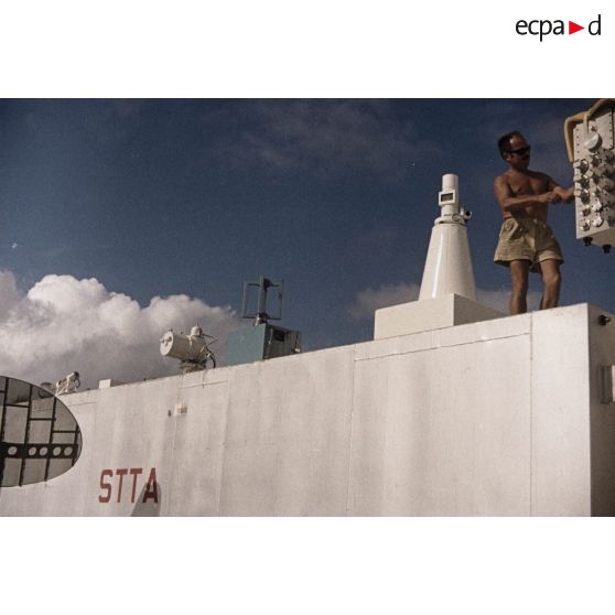 Appareils du Centre d'expérimentations du Pacifique (CEP) sur le toit d'un container portant l'inscription "STTA" et antennes sur l'atoll de Moruroa. Un technicien s'affaire.<br>