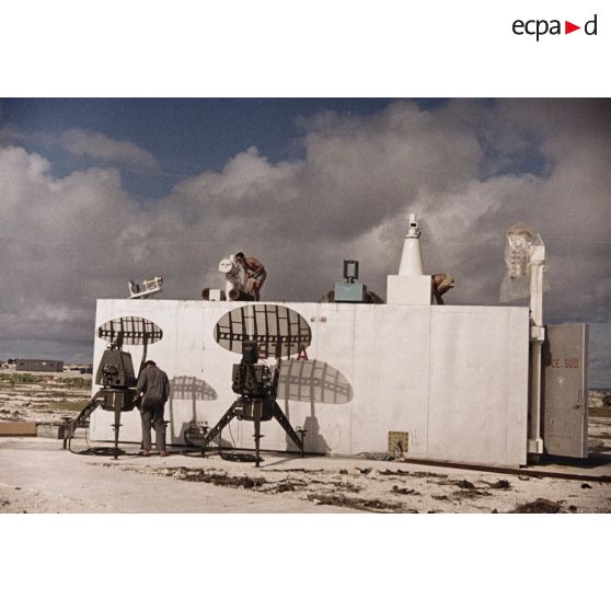 Appareils du Centre d'expérimentations du Pacifique (CEP) sur le toit d'un container portant l'inscription "STTA" et antennes sur l'atoll de Moruroa. Une bâche plastique protège un appareil. Des techniciens s'affairent.