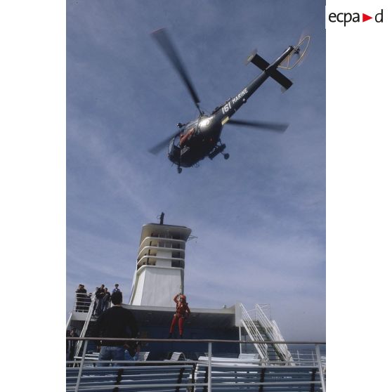 Personnel hélitreuillé sur le pont du ferry Corse depuis l'hélicoptère Alouette-III de l'aéronavale sous le regard de spectateurs.