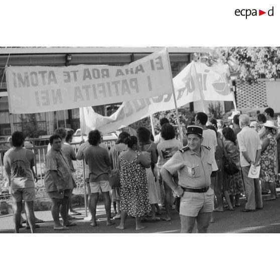 La Gendarmerie nationale à Papeete. Un gendarme devant une manifestation antinucléaire. Banderoles "ei aha roa te atomi i patofita nei" (quel est au juste cet atome qui rayonne ?) et "ia ora te natura" (vive la nature).