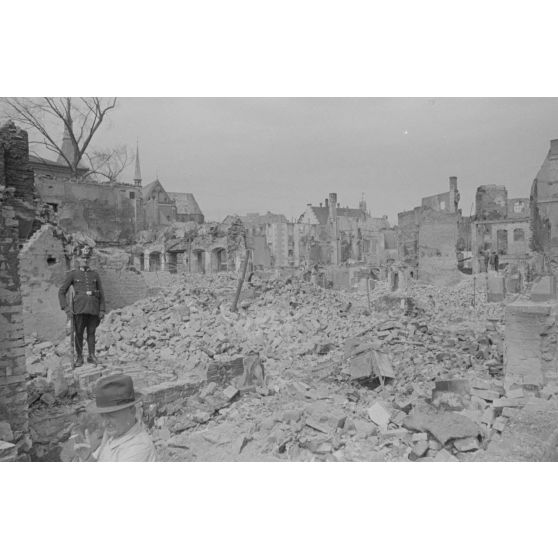 Un policier allemand, tourné vers l’objectif, se tient droit parmi les ruines avec l’église de Sainte-Marie de Rostock derrière.