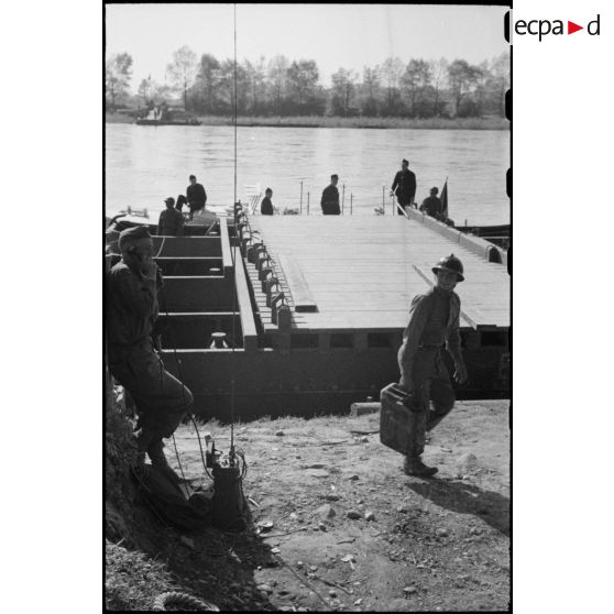 Une portière, constituée de bateaux d'assaut M2 assemblés et manoeuvrés par les sapeurs du Génie, est accostée le long de la berge d'un fleuve.