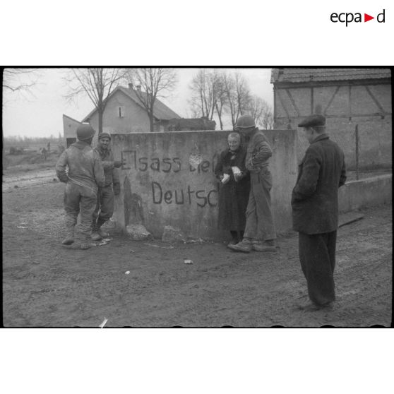 Une habitante du village de Wolfgantzen (Haut-Rhin) à peine sortie de sa cave où elle s'était réfugiée pendant l'attaque du village, donne du pain aux soldats. idem Terre 10093 R10. [description provisoire]