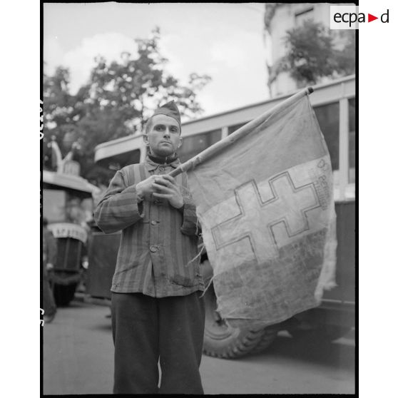 Un déporté politique présente un drapeau à croix de Lorraine sur lequel sont inscrits des noms de camps.