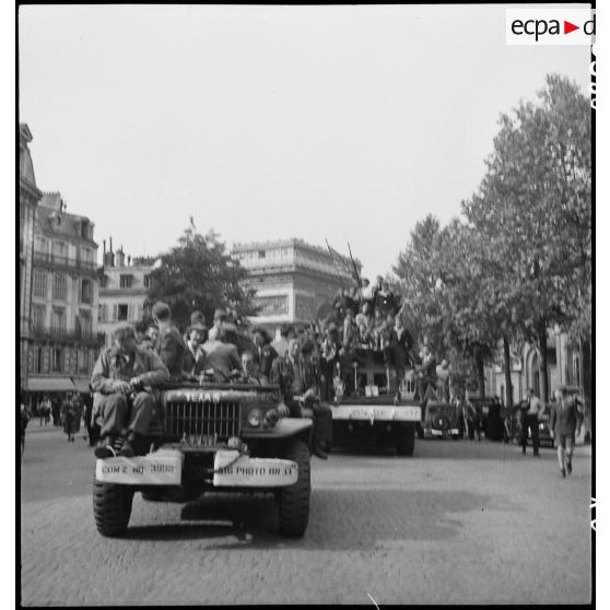 Population civile et militaires français et américains embarqués sur des véhicules.