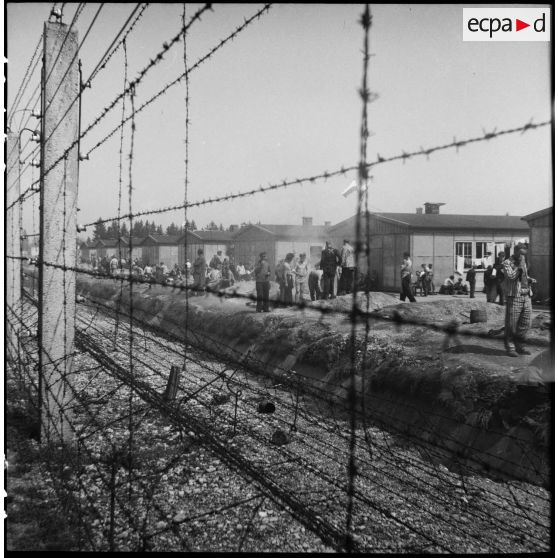 Dans le camp de concentration de Dachau libéré, des déportés rescapés attendent derrière les barbelés, près des baraquements.