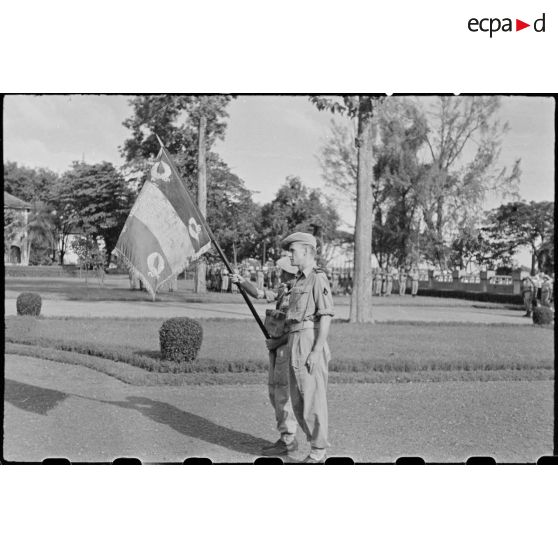 Deux soldats français dont l' un au garde-à-vous et l' autre tenant le drapeau du régiment des tirailleurs cambodgiens. Ce drapeau doit son salut à l' initiative du soldat BOMEAUD qui l' enterra près de TAY-NINH lors d' un combat avec les japonais et repartit plus tard avec un commando pour le déterrer.
