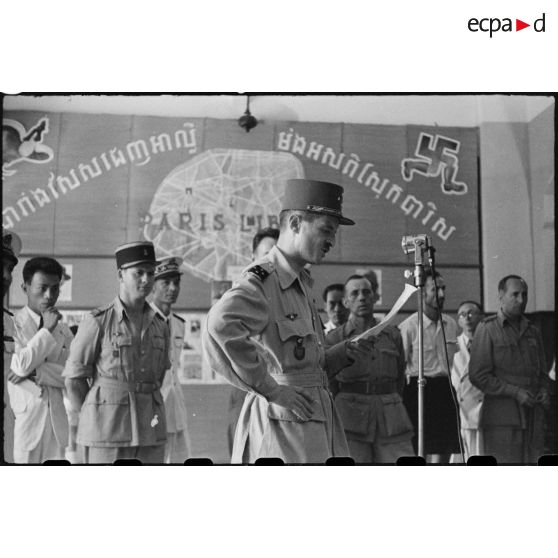 ALLOCUTION . Le général LECLERC (devant un micro) fait un discours dans le hall d' information de PHNOM-PENH sur les liens unissant la FRANCE au CAMBODGE. A l' arrière plan, sur une affiche une caricature de la croix gammée.