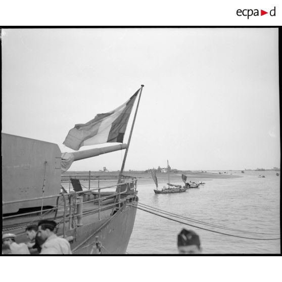 Le drapeau français flotte au milieu du port ; à côté, de nombreuses jonques circulent au milieu des navires.