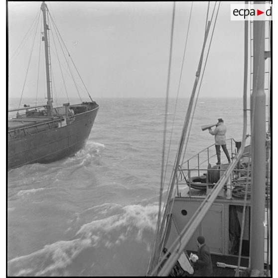 A bord d'un chalutier, un marin hèle au porte-voix l'équipage d'un cargo qui croise sa route.