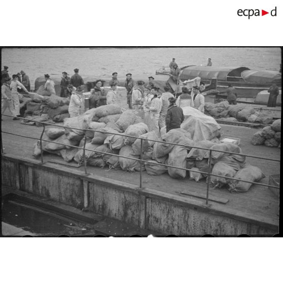Des sacs de vivres, destinés aux navires d'une escadre, sont stockés sur un quai avant d'être embarqués dans des chaloupes ou vedettes.