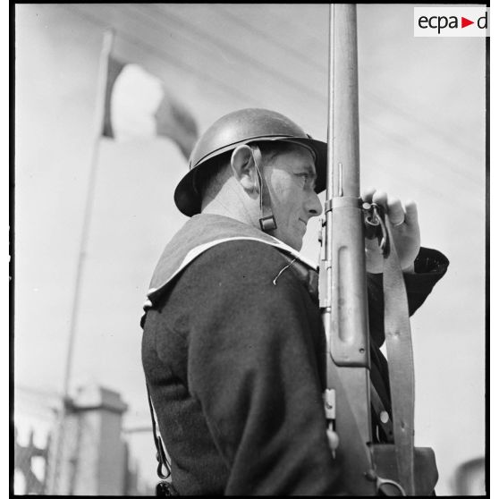 Portrait de profil d'un marin breveté, sentinelle sur un poste de défense côtière, qui présente les armes.