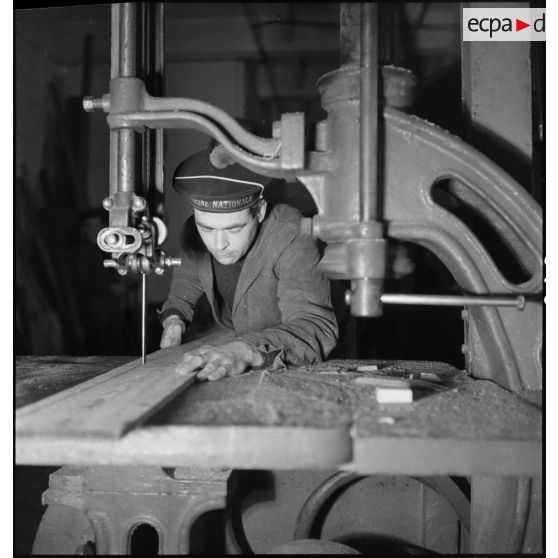 Atelier central de la flotte : un marin utilise une scie à ruban sur une pièce de bois.