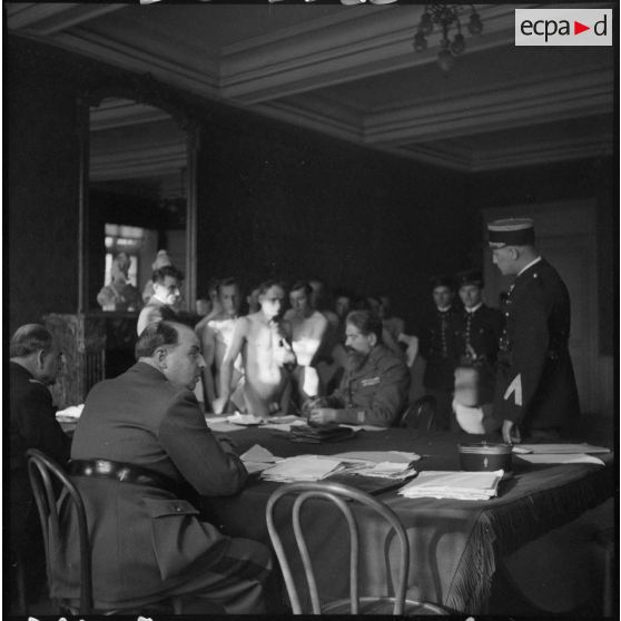 Des hommes nus qui passent en conseil de révision attendent dans une pièce près d'un bureau où se tient un officier.