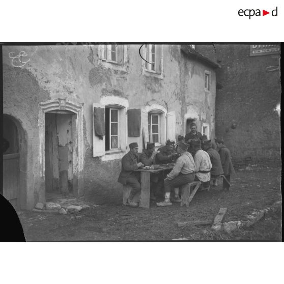 Attablés, des soldats mangent dans la cour d'une ferme.