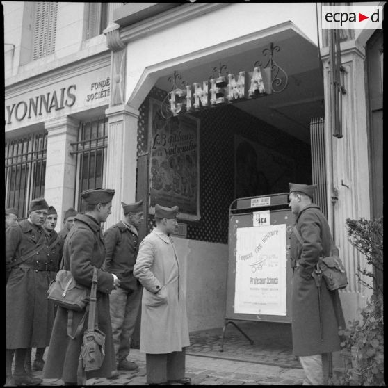 Plan général de l'entrée d'un cinéma, des gens lisent une affiche de cinéma.