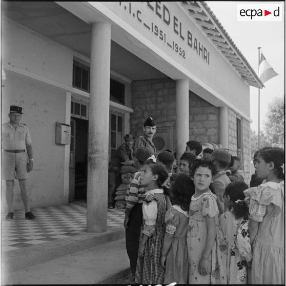 Jeunes élèves algériens en rang devant l'école d'Ouled-El-Bahri sous le regard de rappelés du 20e RD (régiment de dragons).