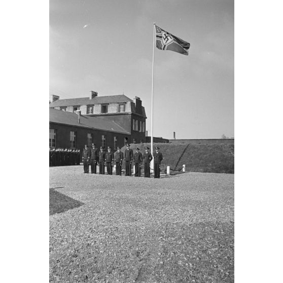 Lors d'une cérémonie de la Kriegsmarine à Saint-Nazaire, des sous-officiers se tiennent au garde-à-vous devant le mat des couleurs orné du drapeau à croix gammée.
