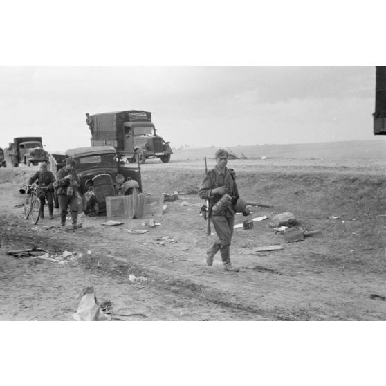 Des fantassins viennent de recevoir leur repas de midi et poursuivent l'avancée parallèlement à des camions Opel Blitz.