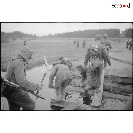 Alors que villageois requis réparent un pont de bois, des fantassins allemands le traversent.