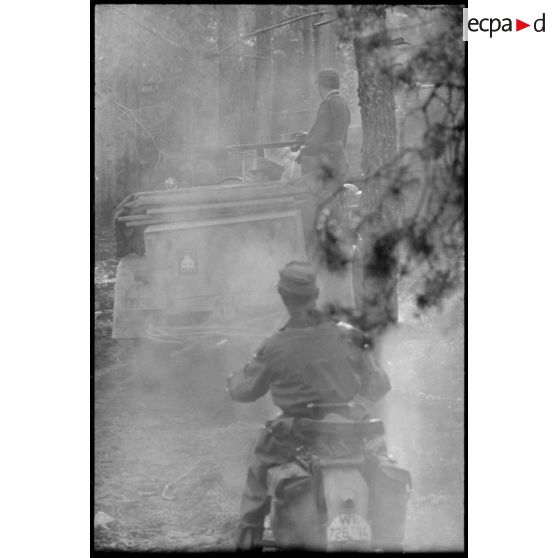 Une colonne de véhicules allemands progresse dans une forêt, au premier plan une moto de la Luftwaffe puis un véhicule sur lequel un insigne d'unité est peint.