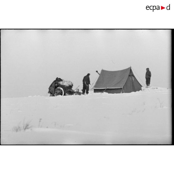 Un abri de fortune dans une tente équipée d'un poêle, une remorque (Infanteriekarren) est recouverte de neige.