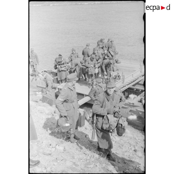 Après la traversée d'une rivière sur un ponton (Brückengerät), le personnel gagne la berge, tandis que des sacs postaux vont être déchargés.