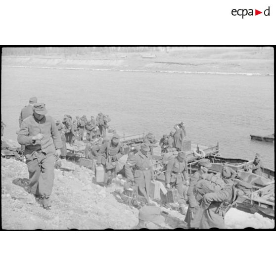 Après la traversée d'une rivière sur un ponton (Brückengerät), le personnel gagne la berge, tandis que des sacs postaux vont être déchargés.