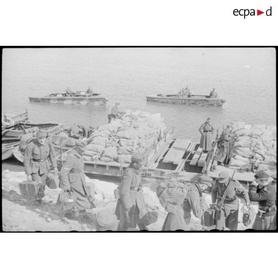 Après la traversée d'une rivière sur un ponton (Brückengerät), le personnel gagne la berge, tandis que des sacs postaux vont être déchargés.