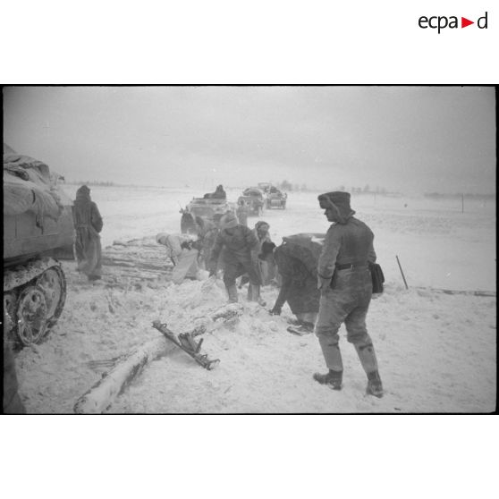 Un semi-chenillé a versé d'un pont de bois, des fantassins tentent de consolider le pont pour le passage d'autres véhicules.