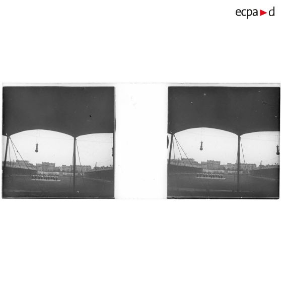 [Spectacle équestre sur un terrain rectangulaire bordé de tribunes, rangée d'immeubles au second plan].<br> [Légende d'origine]