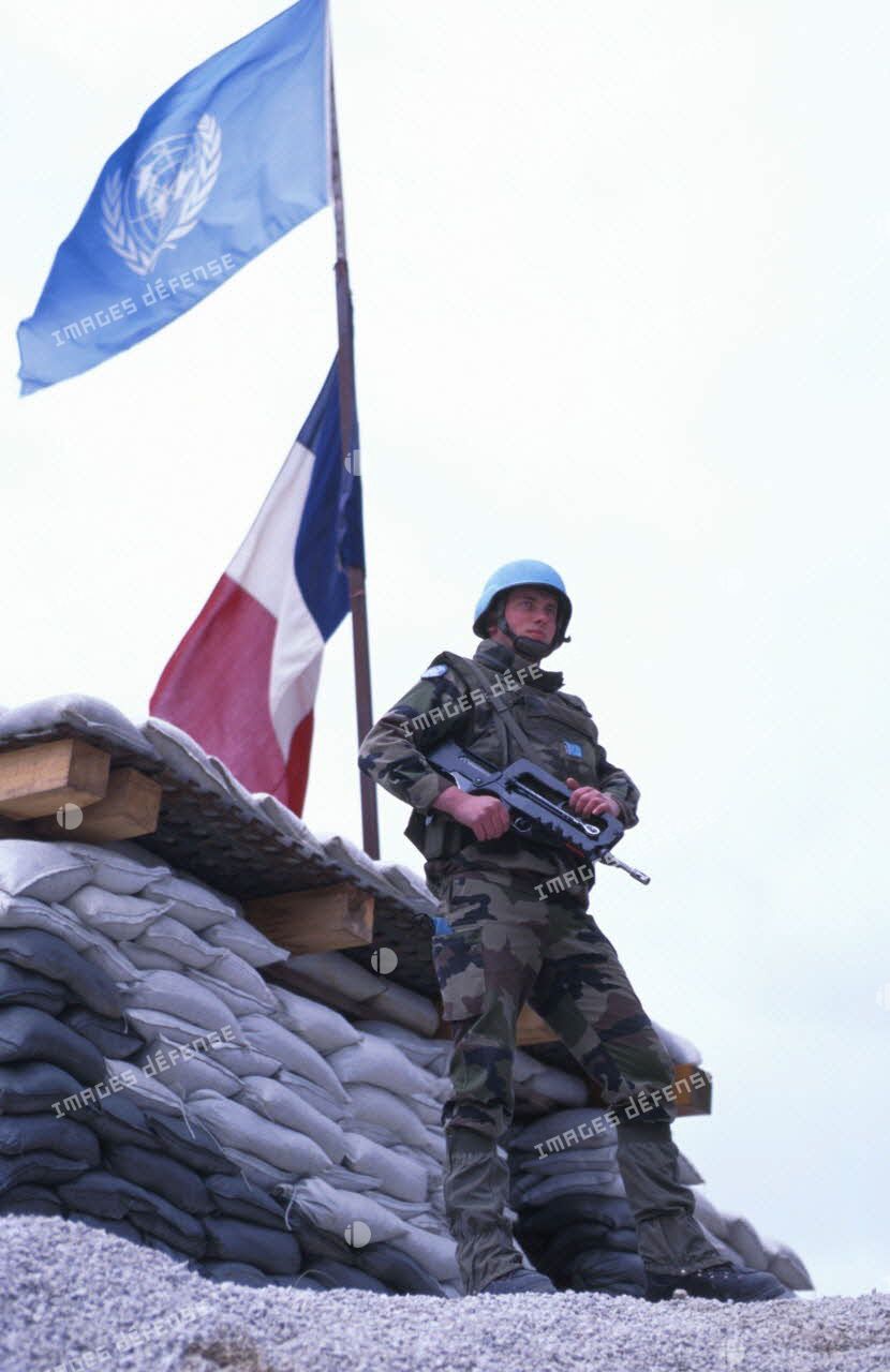 Une sentinelle française pose devant le drapeau de l'ONU et le drapeau français.