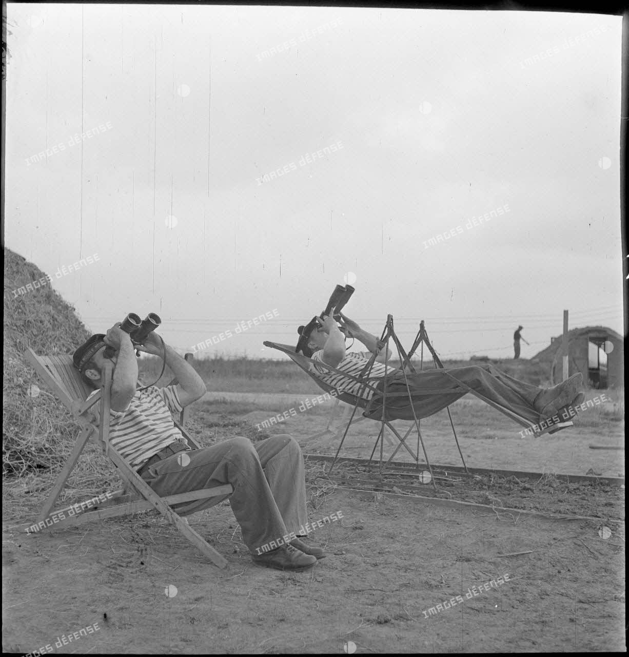 Des marins, appartenant à la 4e batterie de 90 mm contre avions, batterie mobile de défense antiaérienne de la Marine, observent le ciel à l'aide de jumelles, installés dans des chaises longues ou des sièges suspendus.