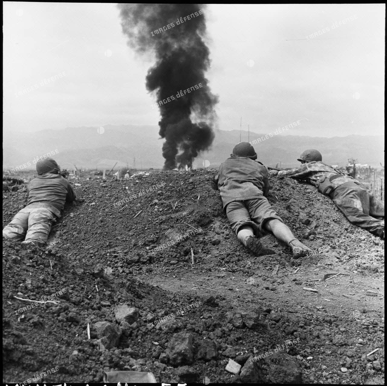 Des soldats en position de tir abrités derrière un monticule de terre alors qu'une épaisse fumée se dégage du terrain d'aviation de Diên Biên Phu.