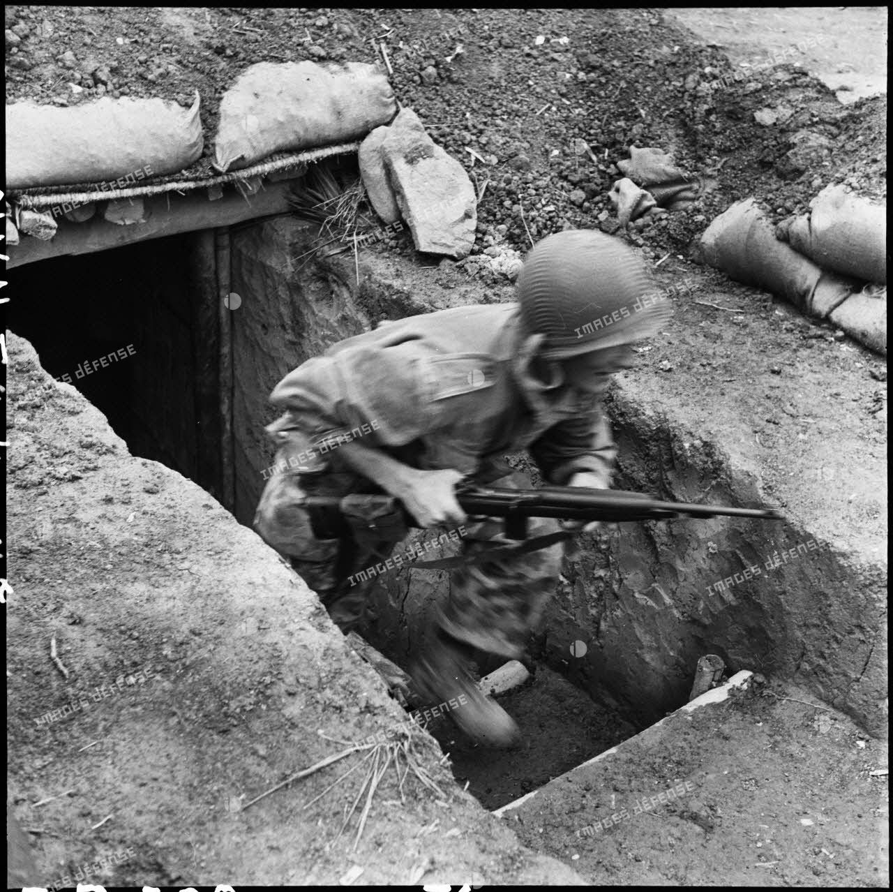 Sous les tirs d'artillerie Viêt-minh, un soldat armée d'une carabine bondit d'un abri pour aller occuper sa position de combat.